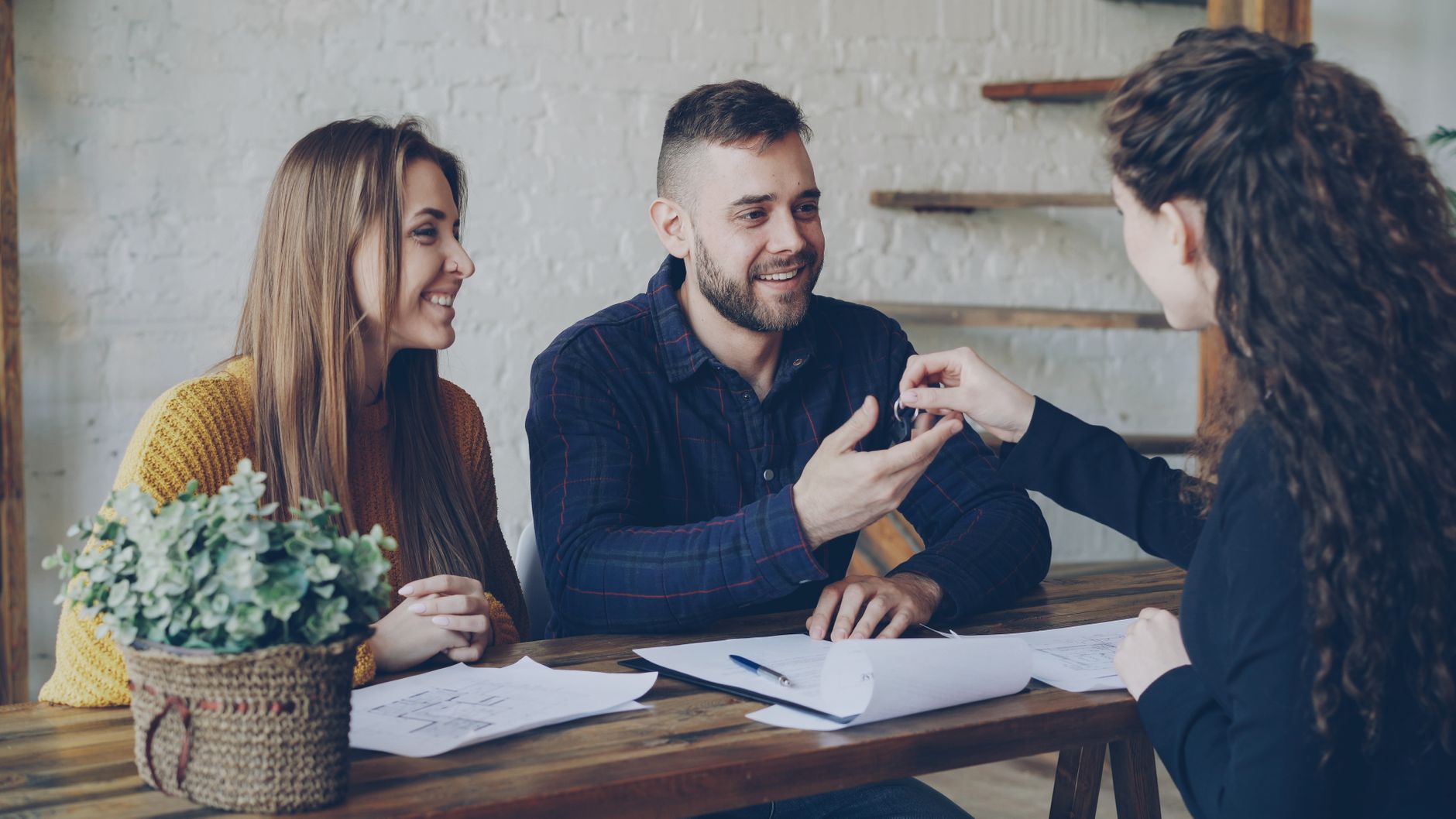 Smiling couple receiving house keys from a female real estate agent at a wooden desk.